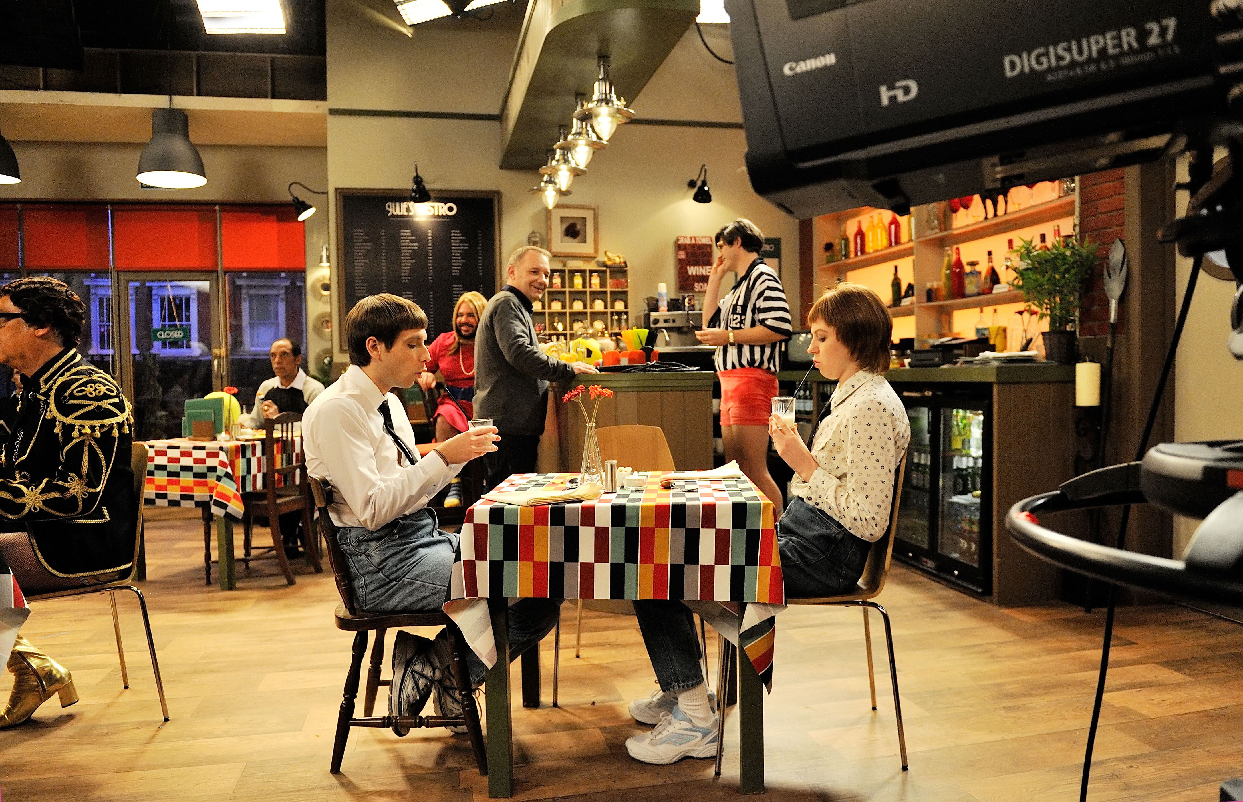 Two actors sit and drink milkshakes on either side of a table. Other cast members are visible in the back of the shot. In front of them all is a television camera.