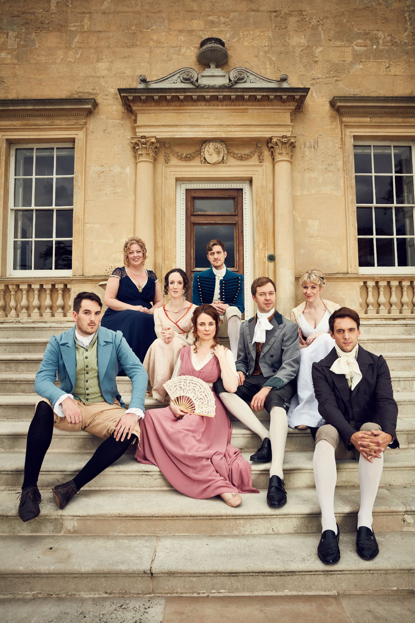 Eight performers in regency costumes sit on the steps of a large manorhouse.