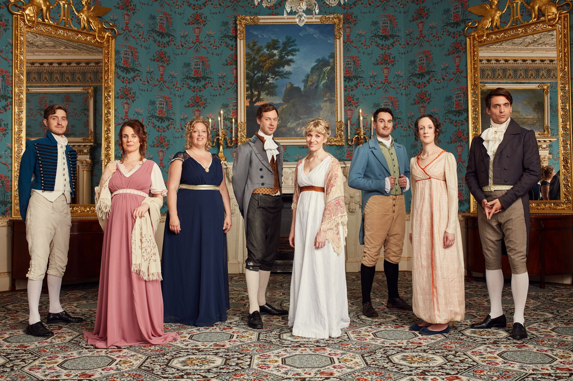 Eight performers in regency costumes line up in front of a painting in a large manor house.