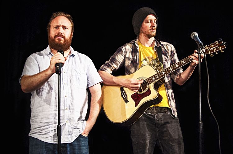 Jonny Donahoe holds a microphone and looks into the camera while Paddy Gervers plays guitar