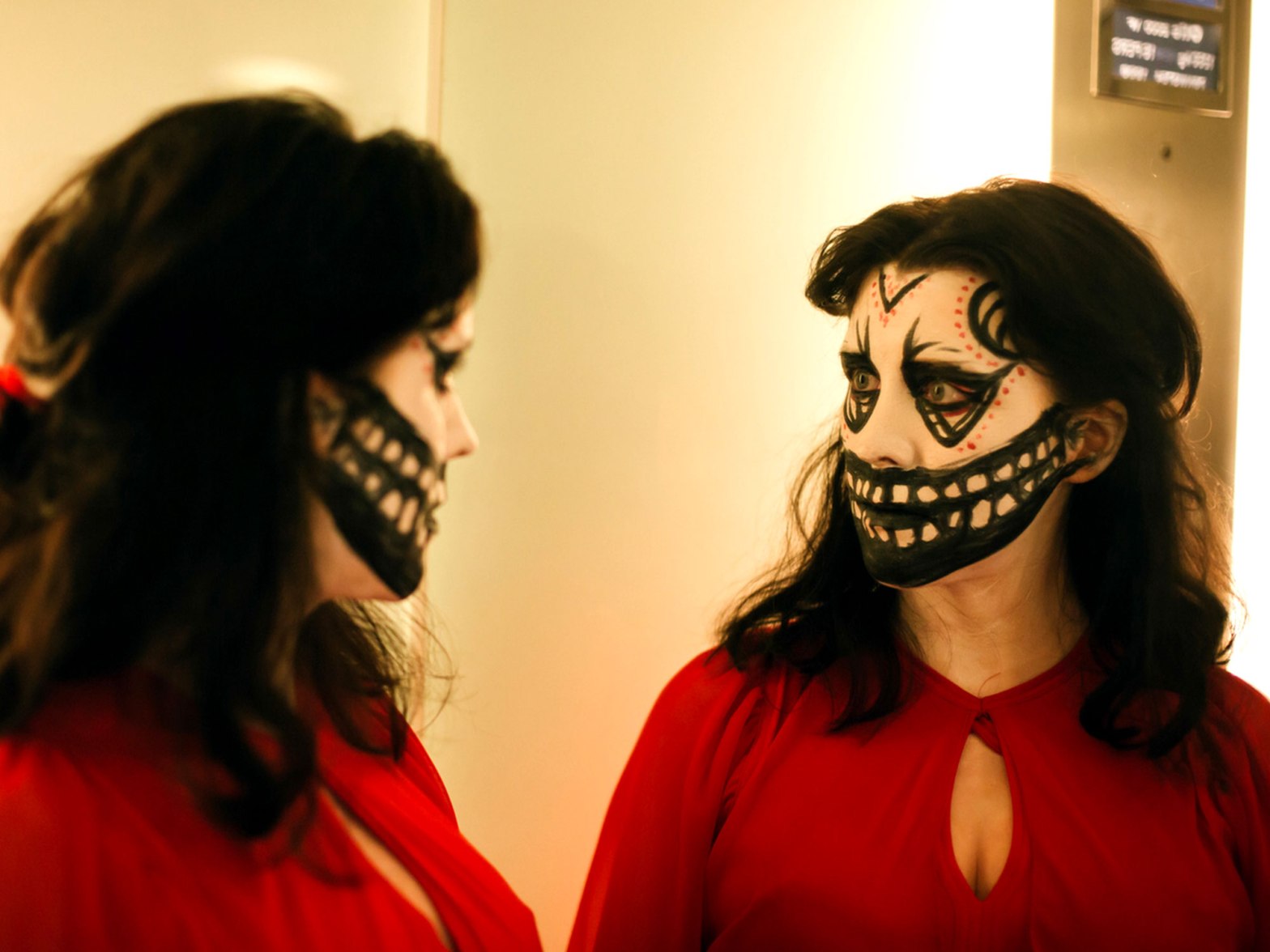 A woman wearing a red dress and skull face-paint, looks at herself in the mirror.