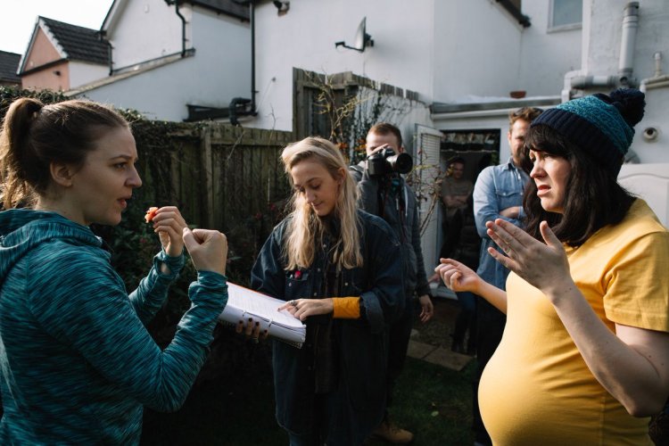 A woman in a blue hoodie holds up her fists in a boxing pose. A pregnant woman opposite her directs her, while another woman stands between them pointing to a page in a script.