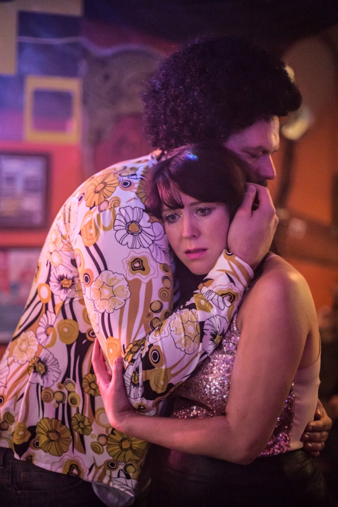 A man wearing an Afro wig and a florid 70s shirt holds a woman tightly to his chest. She is looking sadly away from the scene.