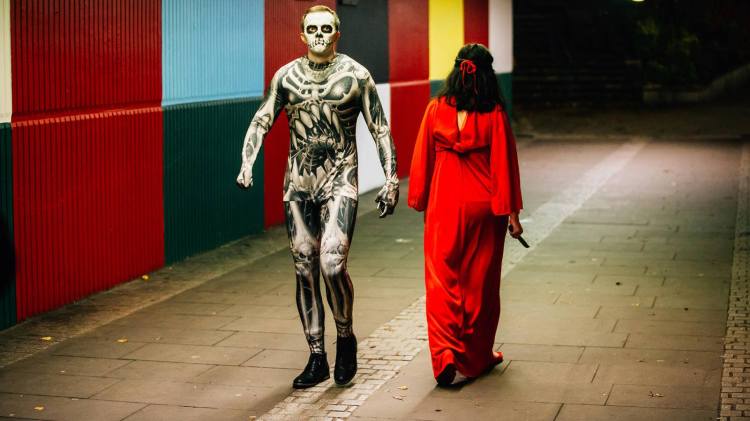 A woman in a red dress holding a knife walks past a man in a skeleton skin-suit in a subway.