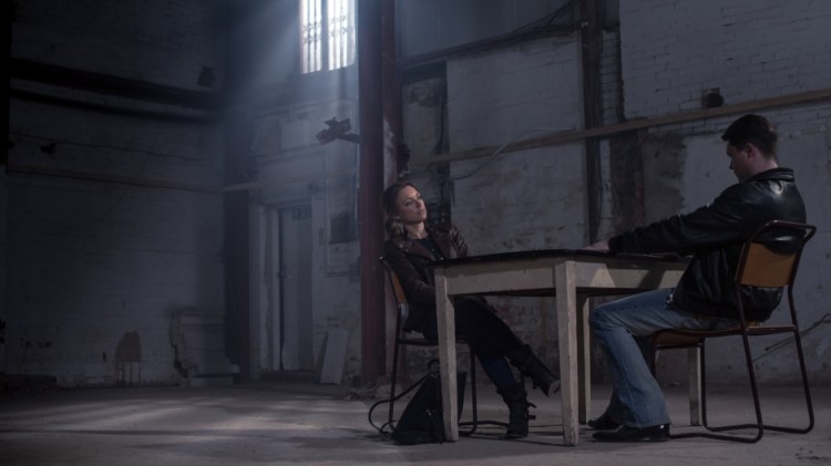 Sarah Alexander and Daniel Rigby sit either side of a table in a large warehouse.
